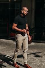 man in black polo shirt and blue denim jeans standing on sidewalk during daytime