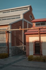 A brick industrial building with metal roofing and piping extends across the structure. The red and beige bricks create a textured appearance, highlighted by sunlight. A circular window is visible on the upper part of one wall, while a barred window is on the lower level surrounded by foliage.