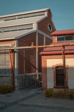 A brick industrial building with metal roofing and piping extends across the structure. The red and beige bricks create a textured appearance, highlighted by sunlight. A circular window is visible on the upper part of one wall, while a barred window is on the lower level surrounded by foliage.