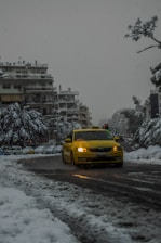 A yellow taxi navigates through a snow-covered city street, surrounded by buildings with snow-laden rooftops. The trees and parked cars are also covered in snow, creating a wintry urban scene. Traffic lights are faintly visible in the background.