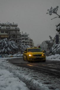 A yellow taxi navigates through a snow-covered city street, surrounded by buildings with snow-laden rooftops. The trees and parked cars are also covered in snow, creating a wintry urban scene. Traffic lights are faintly visible in the background.