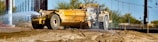 A large yellow construction vehicle sprays water onto a dirt surface, likely to control dust at a construction site. The scene includes dry soil, some sparse vegetation in the background, and power lines overhead.