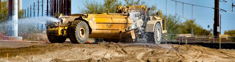 A large yellow construction vehicle sprays water onto a dirt surface, likely to control dust at a construction site. The scene includes dry soil, some sparse vegetation in the background, and power lines overhead.