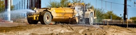 A large yellow construction vehicle sprays water onto a dirt surface, likely to control dust at a construction site. The scene includes dry soil, some sparse vegetation in the background, and power lines overhead.