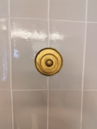 A close-up view of a white tiled wall with a brass circular fixture mounted at the center. The fixture has a smooth, glossy appearance, with concentric circular patterns. The tiles are neatly lined up in a grid pattern and exhibit a slight reflection.