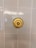 A close-up view of a white tiled wall with a brass circular fixture mounted at the center. The fixture has a smooth, glossy appearance, with concentric circular patterns. The tiles are neatly lined up in a grid pattern and exhibit a slight reflection.