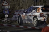 A rally car navigates a turn on a road surrounded by caution tape and road signs, including one warning of deer and another indicating a restricted zone. The vehicle is marked with various sponsorship logos and numbers, emphasizing its participation in a competitive race.