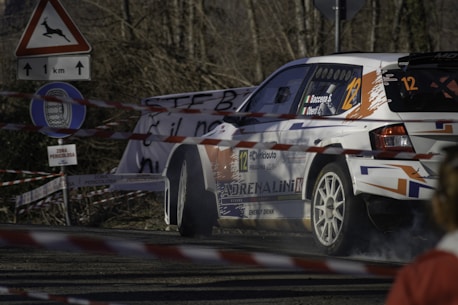 A rally car navigates a turn on a road surrounded by caution tape and road signs, including one warning of deer and another indicating a restricted zone. The vehicle is marked with various sponsorship logos and numbers, emphasizing its participation in a competitive race.