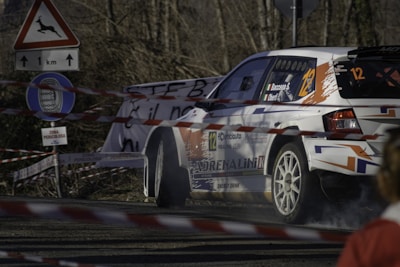 A rally car navigates a turn on a road surrounded by caution tape and road signs, including one warning of deer and another indicating a restricted zone. The vehicle is marked with various sponsorship logos and numbers, emphasizing its participation in a competitive race.