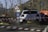 A white rally car with various sponsor logos is making its way through a dirt road or track surrounded by trees without leaves. There are road signs ahead indicating wildlife crossing and a roundabout. Red and white striped tape marks the area, signaling a possible event or restricted zone. A person in a reflective jacket is partially visible, suggesting event supervision or involvement.