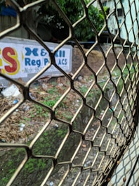 A metal grating or fence is prominently in the foreground, dividing the viewer from a drainage ditch or canal running alongside a wall with a painted advertisement. The ground is covered in grass, leaves, and litter. In the background, partially obscured by foliage, is a white building.