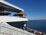 A modern building with an open terrace featuring a thatched roof area overlooks a vast expanse of blue ocean under a clear blue sky. People are dining on the lower terrace, enjoying the scenic view. A staircase with an artistic, geometric design is visible in the foreground.