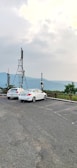 Two white cars are parked in an open lot with satellite dishes mounted on poles nearby. The background features a scenic view of distant hills and a cloudy sky, suggesting an elevated location or lookout point.