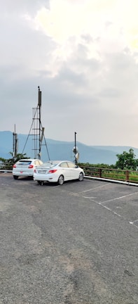 Two white cars are parked in an open lot with satellite dishes mounted on poles nearby. The background features a scenic view of distant hills and a cloudy sky, suggesting an elevated location or lookout point.
