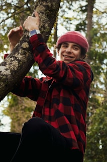 A person wearing a red and black checkered shirt and a red knit cap is climbing a tree. The person has a smile on their face and is looking at the camera. The background displays a forest with green foliage.