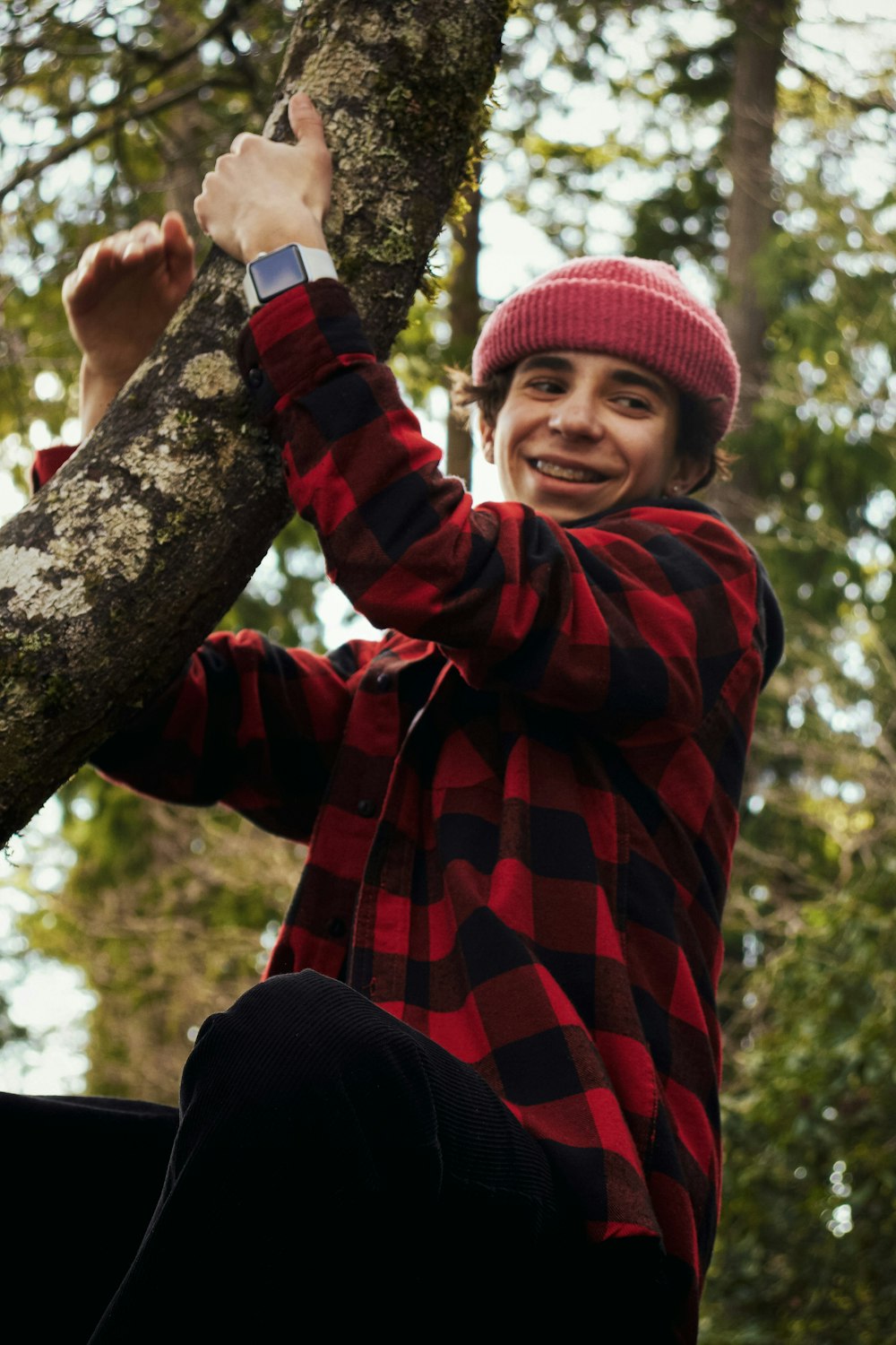 uomo in camicia a quadri rossa e nera e pantaloni neri seduto sul ramo dell'albero durante