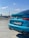 A bright blue BMW M235i is parked on what appears to be a rooftop or parking lot, with a clear blue sky and a television tower in the background. The car's license plate is visible, and the area appears calm and sunny.