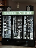 A wine refrigerator with three sections, each filled with various bottles of wine and spirits. The top portion displays the phrase 'Truth in Dirt' in illuminated letters. Several bottles are lying horizontally on the shelves, showcasing their labels. Glass bottles, likely empty, are also visible at the base. The fridge is set against a dark background with additional wine bottles stored on shelves to the left.