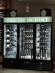 A wine refrigerator with three sections, each filled with various bottles of wine and spirits. The top portion displays the phrase 'Truth in Dirt' in illuminated letters. Several bottles are lying horizontally on the shelves, showcasing their labels. Glass bottles, likely empty, are also visible at the base. The fridge is set against a dark background with additional wine bottles stored on shelves to the left.