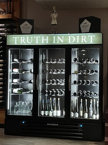A wine refrigerator with three sections, each filled with various bottles of wine and spirits. The top portion displays the phrase 'Truth in Dirt' in illuminated letters. Several bottles are lying horizontally on the shelves, showcasing their labels. Glass bottles, likely empty, are also visible at the base. The fridge is set against a dark background with additional wine bottles stored on shelves to the left.