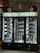 A wine refrigerator with three sections, each filled with various bottles of wine and spirits. The top portion displays the phrase 'Truth in Dirt' in illuminated letters. Several bottles are lying horizontally on the shelves, showcasing their labels. Glass bottles, likely empty, are also visible at the base. The fridge is set against a dark background with additional wine bottles stored on shelves to the left.
