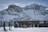 Several people are dressed in winter gear and ski equipment, standing in front of a wooden ski hut labeled 'Pizol Berghotelhütte'. Snow-covered mountains with jagged peaks rise dramatically in the background under a clear blue sky.