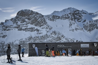 Several people are dressed in winter gear and ski equipment, standing in front of a wooden ski hut labeled 'Pizol Berghotelhütte'. Snow-covered mountains with jagged peaks rise dramatically in the background under a clear blue sky.