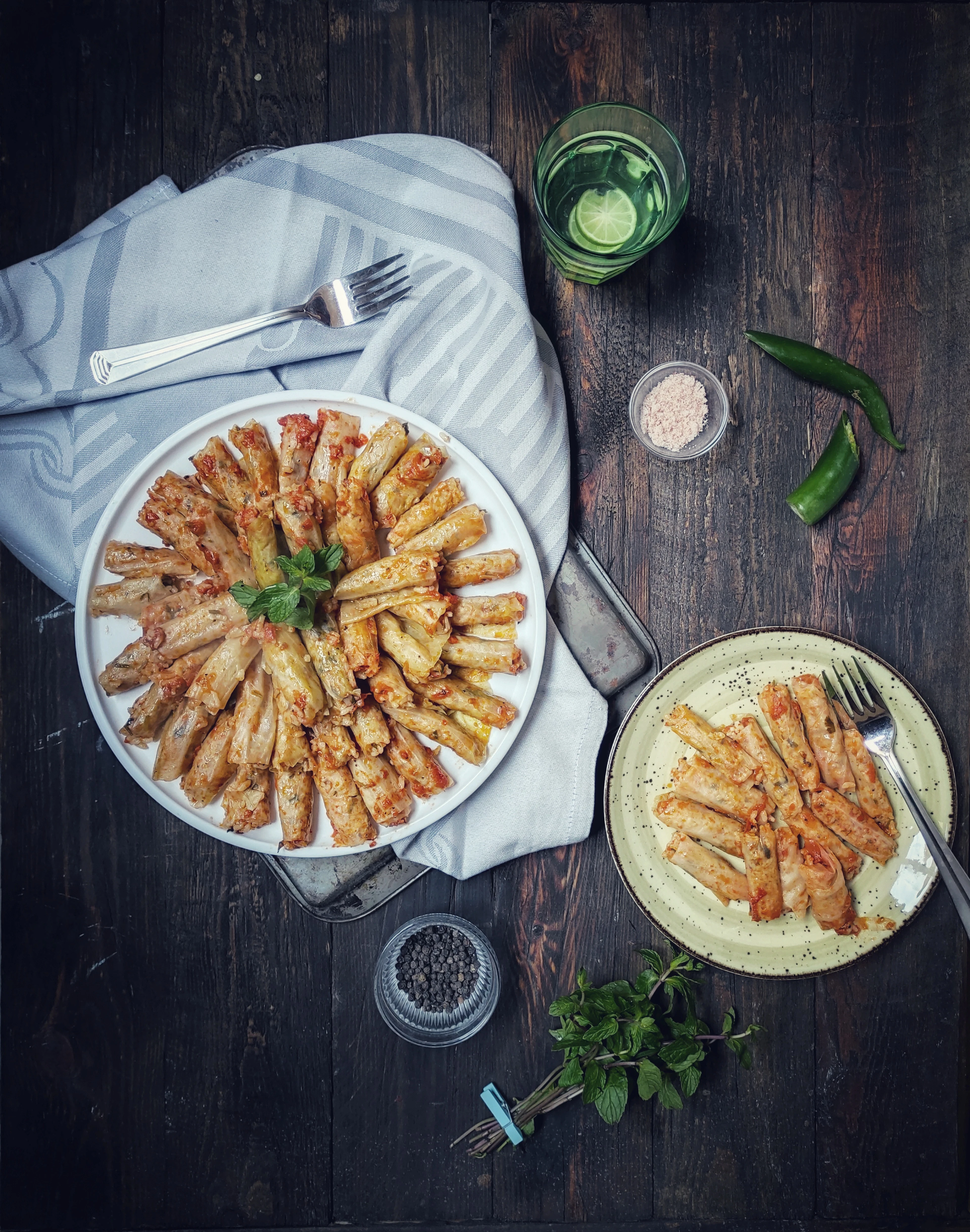 Egyptian mahshi: cabbage leaves rolled around spiced rice and cooked in tomato, fanned in a ring on a white platter with a sprig of mint at the centre, a second plateful, fresh mint and a glass of limed water beside it on dark wood.
