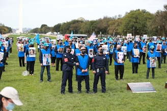 A large group of people wearing blue vests are gathered outdoors, holding photographs of various individuals. Some are wearing masks and hats with a specific emblem. They are on a grassy area, with some people holding flags. In the background, there are trees and a monument.