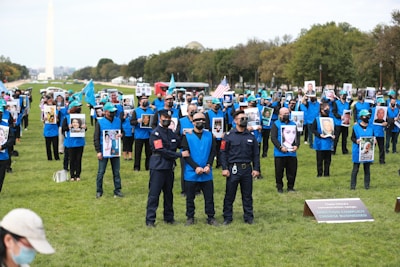 A large group of people wearing blue vests are gathered outdoors, holding photographs of various individuals. Some are wearing masks and hats with a specific emblem. They are on a grassy area, with some people holding flags. In the background, there are trees and a monument.