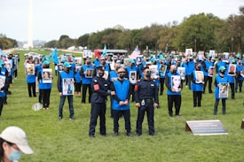 A large group of people wearing blue vests are gathered outdoors, holding photographs of various individuals. Some are wearing masks and hats with a specific emblem. They are on a grassy area, with some people holding flags. In the background, there are trees and a monument.