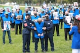 A group of individuals dressed in blue vests, many of whom are holding photos, stand on a grassy field. Two people in uniform are handcuffing a person in the foreground. Several of the individuals wear face masks.