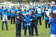 A group of individuals dressed in blue vests, many of whom are holding photos, stand on a grassy field. Two people in uniform are handcuffing a person in the foreground. Several of the individuals wear face masks.