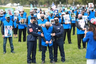 A group of individuals dressed in blue vests, many of whom are holding photos, stand on a grassy field. Two people in uniform are handcuffing a person in the foreground. Several of the individuals wear face masks.