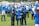 A group of individuals dressed in blue vests, many of whom are holding photos, stand on a grassy field. Two people in uniform are handcuffing a person in the foreground. Several of the individuals wear face masks.