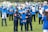 A group of individuals dressed in blue vests, many of whom are holding photos, stand on a grassy field. Two people in uniform are handcuffing a person in the foreground. Several of the individuals wear face masks.