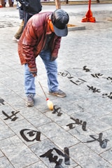An elderly man is engaged in calligraphy, using a large brush to write Chinese characters on a tiled pavement. He is dressed in a brown leather jacket, jeans, and a black cap. The characters appear to be written with water, creating a transient art form on the stone surface.