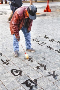 An elderly man is engaged in calligraphy, using a large brush to write Chinese characters on a tiled pavement. He is dressed in a brown leather jacket, jeans, and a black cap. The characters appear to be written with water, creating a transient art form on the stone surface.