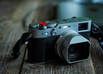 A vintage-style digital camera with a silver metal body and black leather grip rests on a rustic wooden surface. The lens is prominently featured, and a red button stands out on the top of the camera. Soft lighting creates a moody, nostalgic atmosphere.