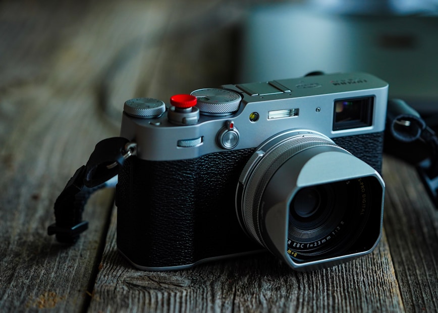 A vintage-style digital camera with a silver metal body and black leather grip rests on a rustic wooden surface. The lens is prominently featured, and a red button stands out on the top of the camera. Soft lighting creates a moody, nostalgic atmosphere.
