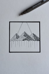 A black ink drawing depicting a stylized mountain landscape within a square frame. The mountains are rendered with intricate linework, and two diagonal lines extend upward from the peaks. A black ink pen is positioned diagonally above the drawing, suggesting a creative process in progress.