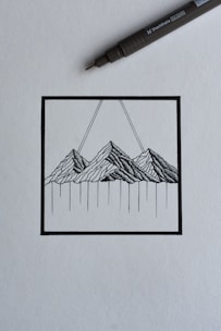 A black ink drawing depicting a stylized mountain landscape within a square frame. The mountains are rendered with intricate linework, and two diagonal lines extend upward from the peaks. A black ink pen is positioned diagonally above the drawing, suggesting a creative process in progress.