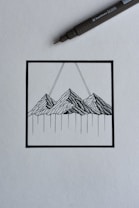 A black ink drawing depicting a stylized mountain landscape within a square frame. The mountains are rendered with intricate linework, and two diagonal lines extend upward from the peaks. A black ink pen is positioned diagonally above the drawing, suggesting a creative process in progress.