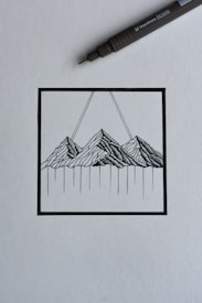 A black ink drawing depicting a stylized mountain landscape within a square frame. The mountains are rendered with intricate linework, and two diagonal lines extend upward from the peaks. A black ink pen is positioned diagonally above the drawing, suggesting a creative process in progress.