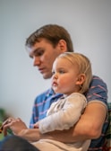 A man is holding a young child on his lap, both appearing to be engaged in an activity. The background is softly blurred, focusing attention on their expressions.