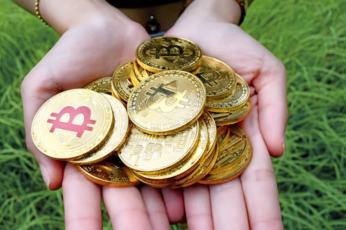 A pair of hands is holding a pile of shiny gold coins featuring the Bitcoin symbol. The background consists of green grass.