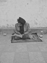 An individual sits cross-legged on a patterned mat against a textured wall. Worn clothing and an uneven surface convey a sense of hardship. A plastic bottle is visible beside the person, and their gaze is directed to the side.