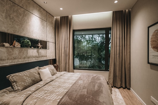 A cozy bedroom features a large bed with patterned bedding and several pillows. The walls are decorated with subtle, textured patterns and a small horizontal shelf holds decorative items and plants. A large window with thick curtains provides natural light and offers a view of greenery outside. The room has a warm, inviting ambiance with soft lighting and neutral tones.