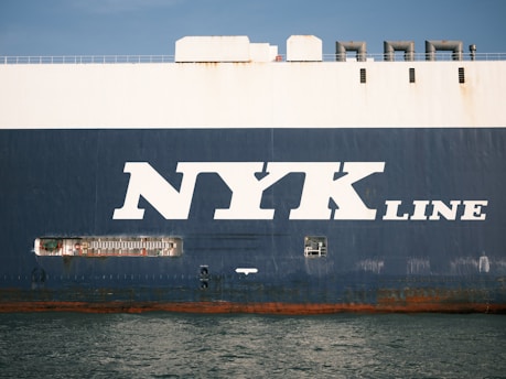 The side view of a large cargo ship with the words 'NYK LINE' prominently displayed in large white letters on a dark blue background. The ocean is visible at the bottom edge of the image, reflecting the ship's hull.