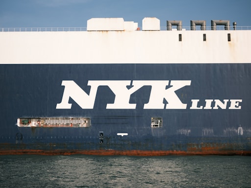 The side view of a large cargo ship with the words 'NYK LINE' prominently displayed in large white letters on a dark blue background. The ocean is visible at the bottom edge of the image, reflecting the ship's hull.